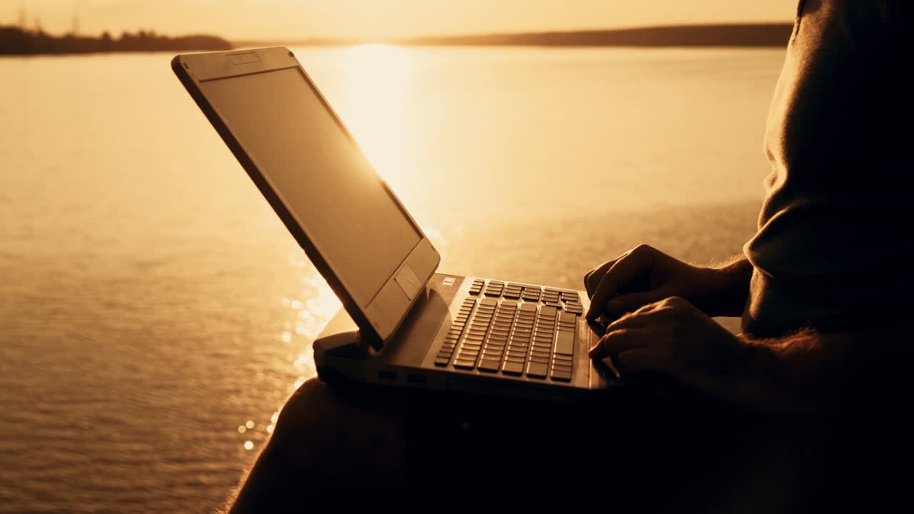 A man is sitting and working on the laptop near the lake in the background of the sunset at the weekend. Close-up
