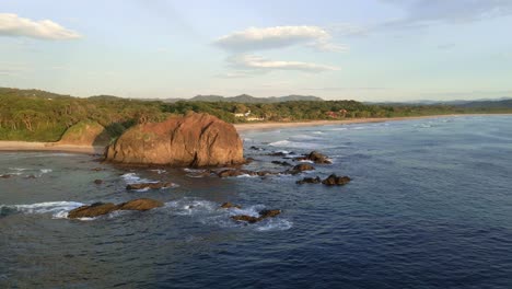 drone approaching a big beach rock then passing it closely and flying over people enjoying the sunset at playa grande