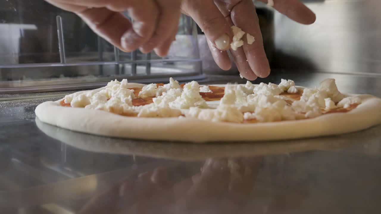 레스토랑 주방 조리대에 있는 피자 도우에 치즈를 뿌린 주방장의 손을 가까이서 볼 수 있습니다.