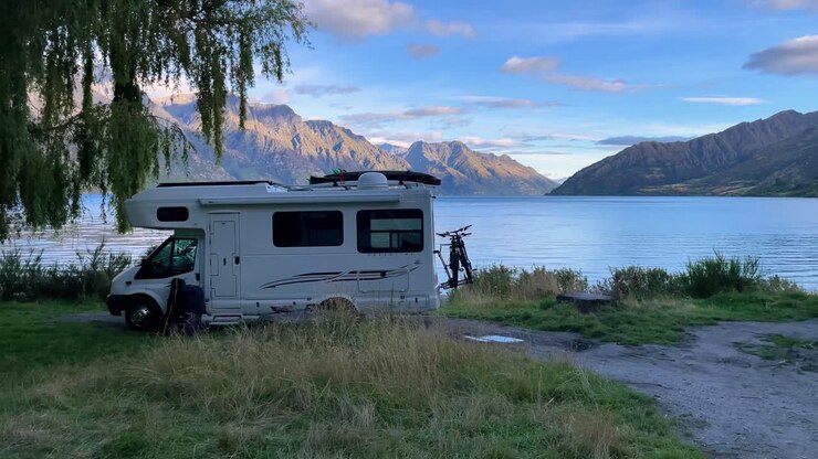 Huzurlu bir yaz akşamında, gün batımında, dağ manzaralı sakin bir gölün önünde park etmiş bir karavan.