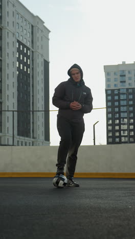 hombre en una arena deportiva urbana se equilibra en una pierna en una pelota de fútbol, manos juntas, girando la cabeza, edificios de gran altura en el fondo bajo un cielo despejado