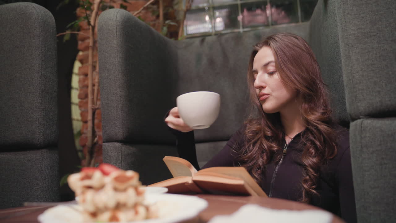 Lady in black sips latte while reading brown book inside cozy booth, gently cleaning mouth with hand, surrounded by soft cushions, waffles on plate, and warm brick interior in relaxed atmosphere