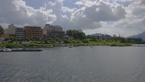 A-picturesque-boat-perspective-captures-the-charm-of-Kawaguchiko-village-in-Japan-on-a-gently-overcast-day,-complemented-by-the-graceful-presence-of-swan-boats