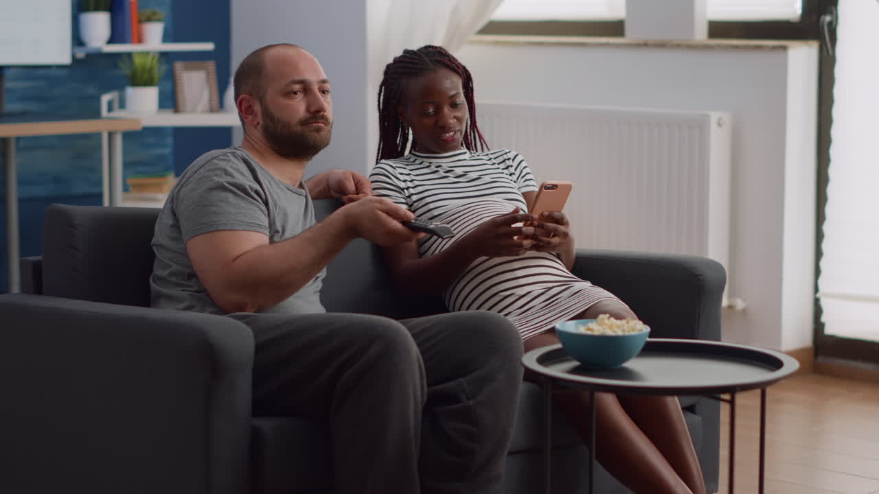Young interracial couple expecting child and sitting on sofa