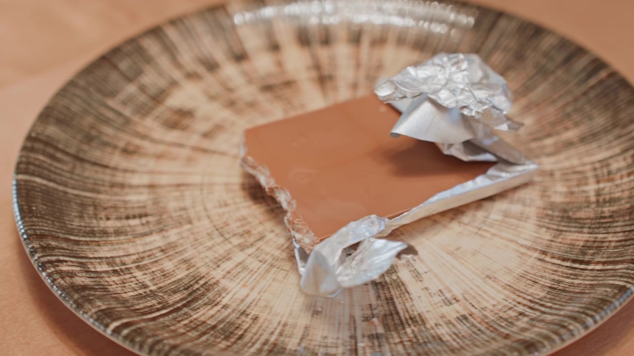 Empty ceramic plate before chocolate appears, hand reaches in to place partially wrapped milk chocolate, foil crumples, textured plate pattern, warm surface, cozy snack preparation, closeup sequence