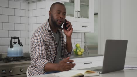 Afroamerikanischer-Mann,-Der-Zu-Hause-In-Der-Küche-Arbeitet-Und-Auf-Dem-Smartphone-Spricht-Und-Einen-Laptop-Benutzt
