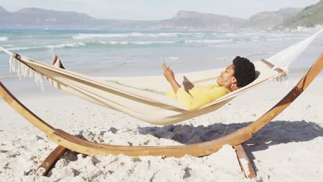 happy african american woman reading and lying in hammock on sunny beach