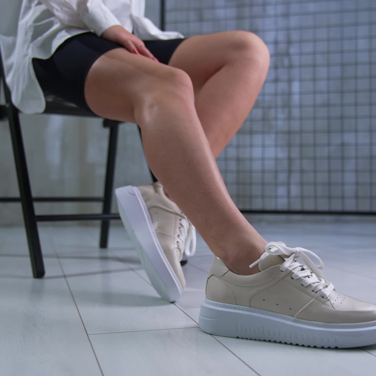 Woman in black bicycle shorts and white shirt sitting on the chair in studio. Lady touches her beige eco leather sneakers. Close up