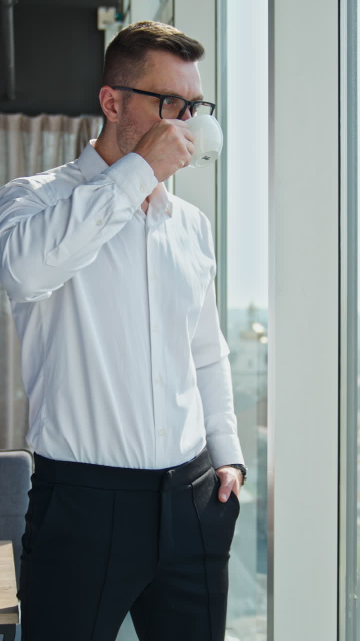 Pensive confident male entrepreneur drinking coffee standing at the window in the office. Businessman having lunch enjoying city view. Vertical video