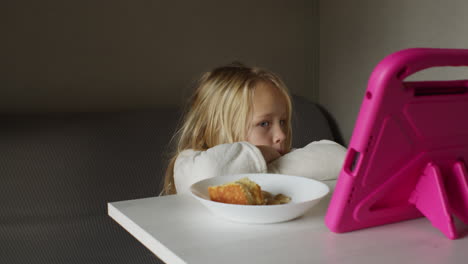 a little girl sitting at a table looking at a tablet while eating breakfast.