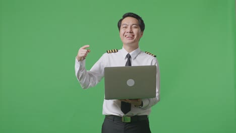 asian man pilot smiling and pointing to a laptop in his hand while standing in the green screen background studio