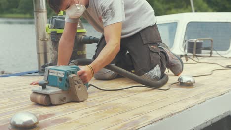 young man starts belt sanding planks on wooden boat forecabin