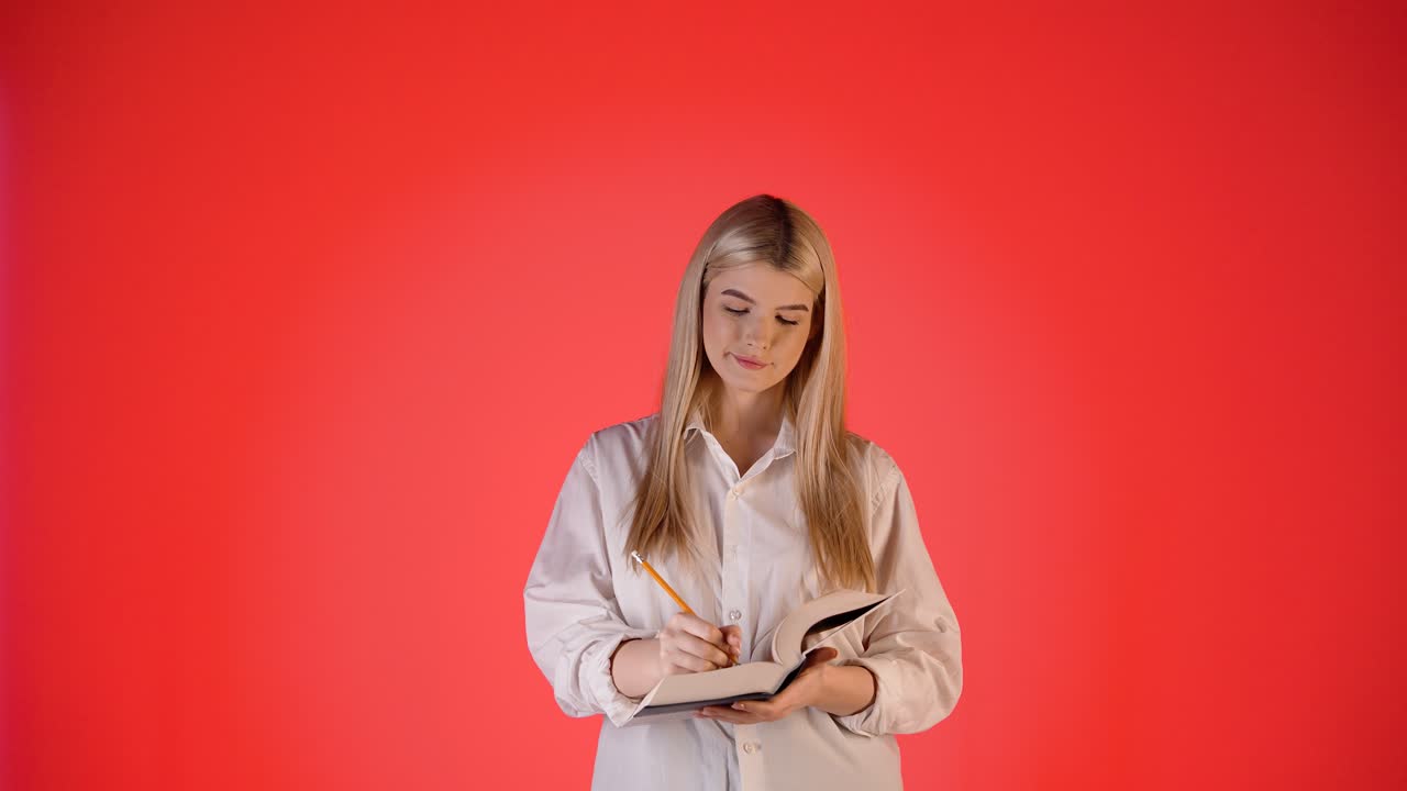 Ung pen kvinne tenker, deretter skriver notater på notisblokk, studiobilde med rød bakgrunn