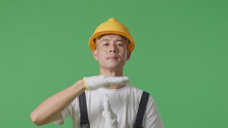 Close Up Of Asian Man Painter Wearing Safety Helmet Looking At Camera And Showing Time Out Hands Gesture While Standing In The Green Screen Background Studio