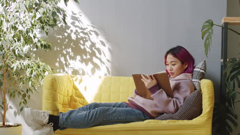 Young-Asian-Woman-Lying-on-Sofa-and-Reading-Book-in-Office