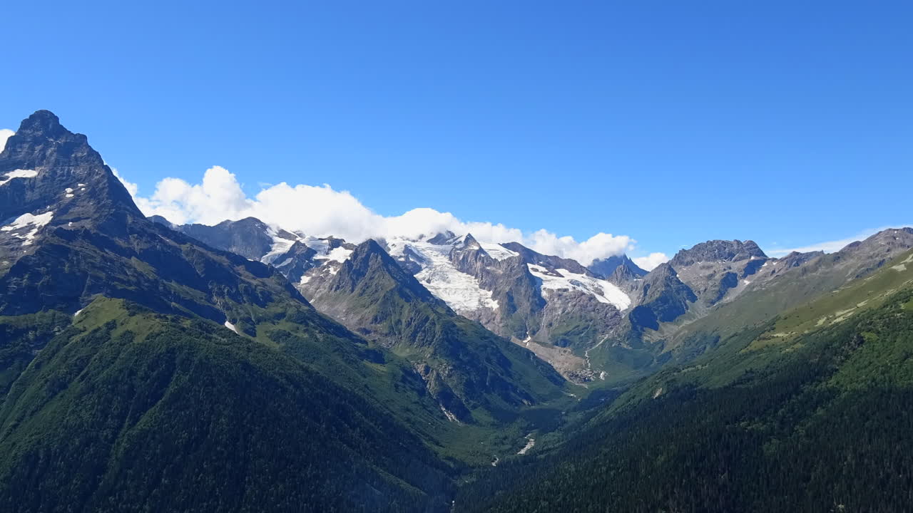 ロシア、コーカサス地方の雪を頂いた山々が連なる緑豊かな山間の谷の上を雲が漂うタイムラプス映像。移り変わる光と劇的な高山風景が映し出されています