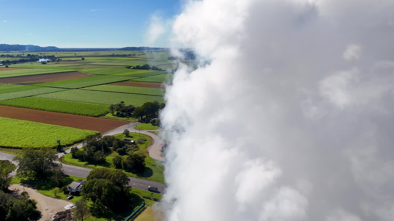Drone footage captures smoke rising from a factory amidst lush farmland under clear blue skies