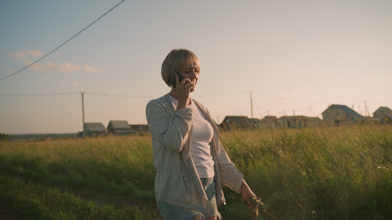 mujer en pantalones cortos de jean caminando en una llamada telefónica a lo largo de un campo de hierba durante el amanecer, con una correa de perro en una mano, el fondo con hierba alta, postes eléctricos y edificios rurales distantes