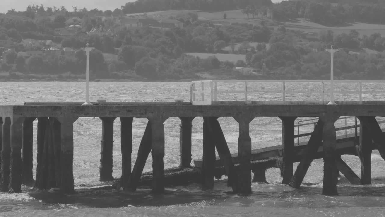 Monochrome wide shot of waves hitting wooden pier, overcast daylight, calm rural waterfront setting