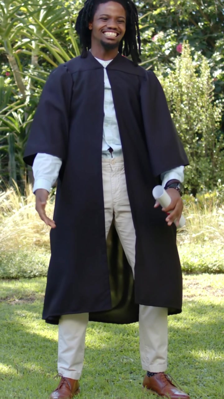 Video verticale: Uomo in toga nera che tiene diploma e tocco, festeggia la laurea alzando le braccia in giardino.
