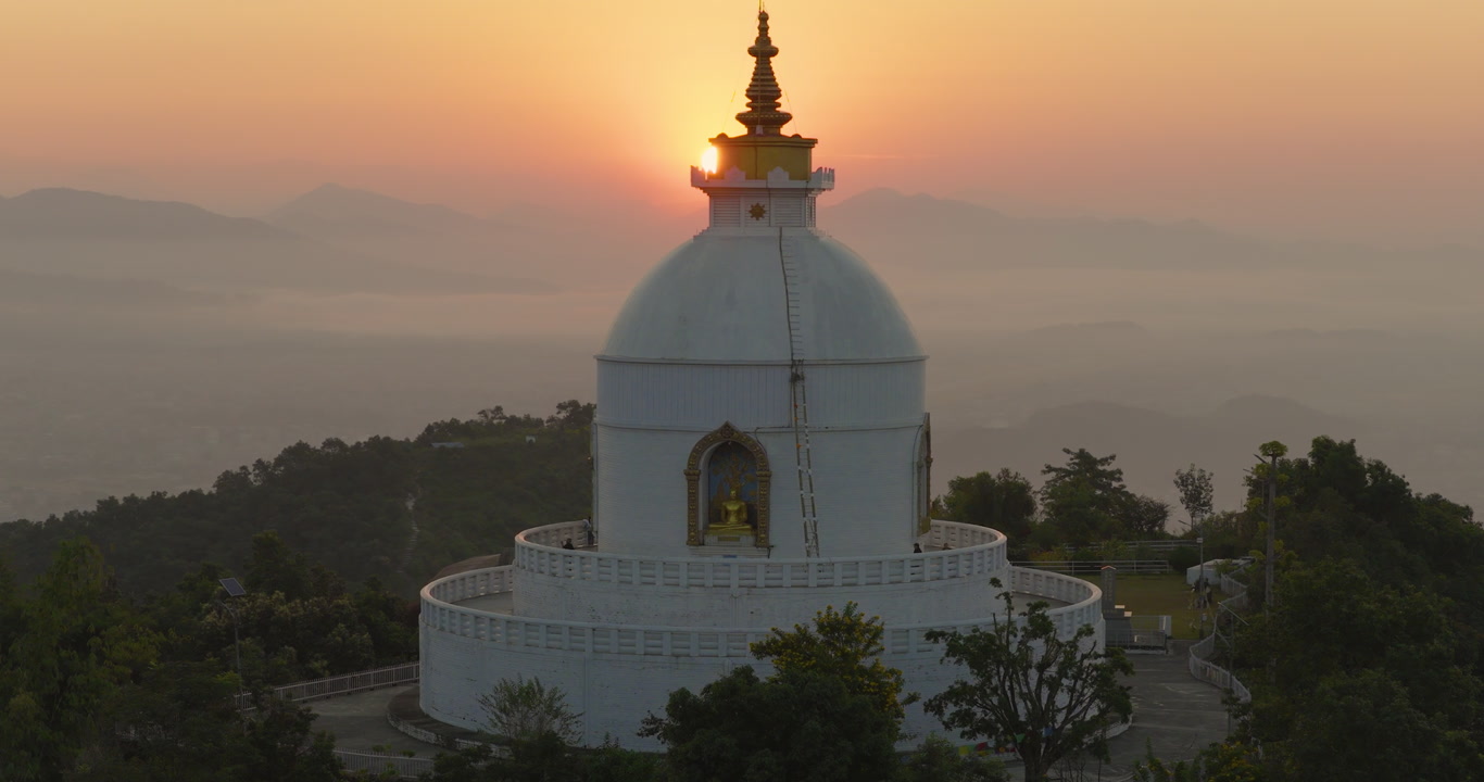 Drone shot of the Buddhist monument World Peace Stupa Pokhara Nepal peak illuminated by golden sunlight beneath an orange sky during sunrise.