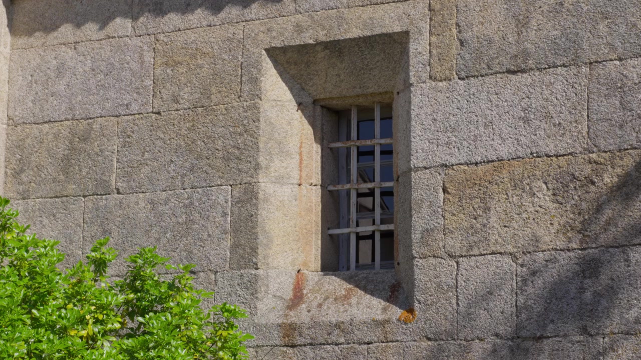 Primo piano dettagliato di un muro di pietra texturizzato con una finestra con sbarre di ferro, immerso nella luce solare naturale, con vegetazione in primo piano.