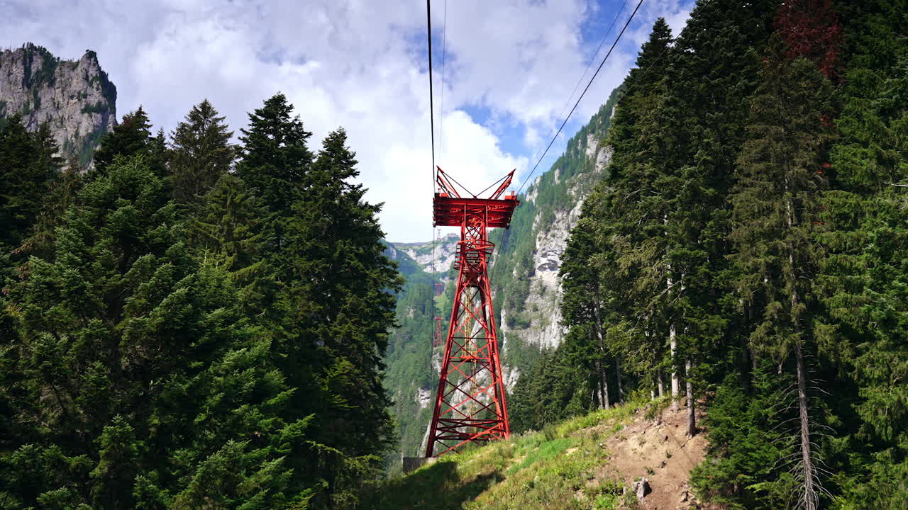 Cable car in the mountains. A striking red cable car tower stands among dense green trees with towering mountains in the background on a clear day