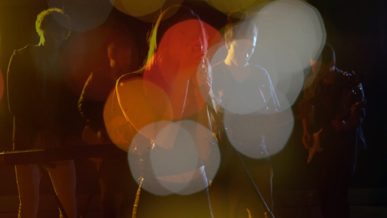 Music band performing on stage with female vocalist holding mic, showing bokeh light effects