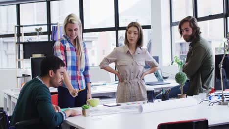 Diverse-office-colleagues-wearing-face-masks-discussing-over-blueprint-plan-at-modern-office