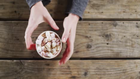 overhead view of hands holding a hot chocolate with marshmallows against wooden surface