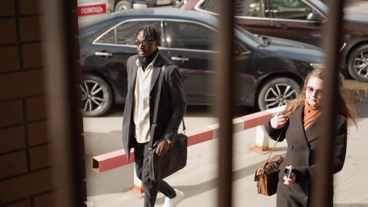 Two professionals walk by entrance, modern young lad looks in woman direction, sunlight on coats, iron bars foreground, parked cars background, casual city commute, crosswalk vibe in bright afternoon