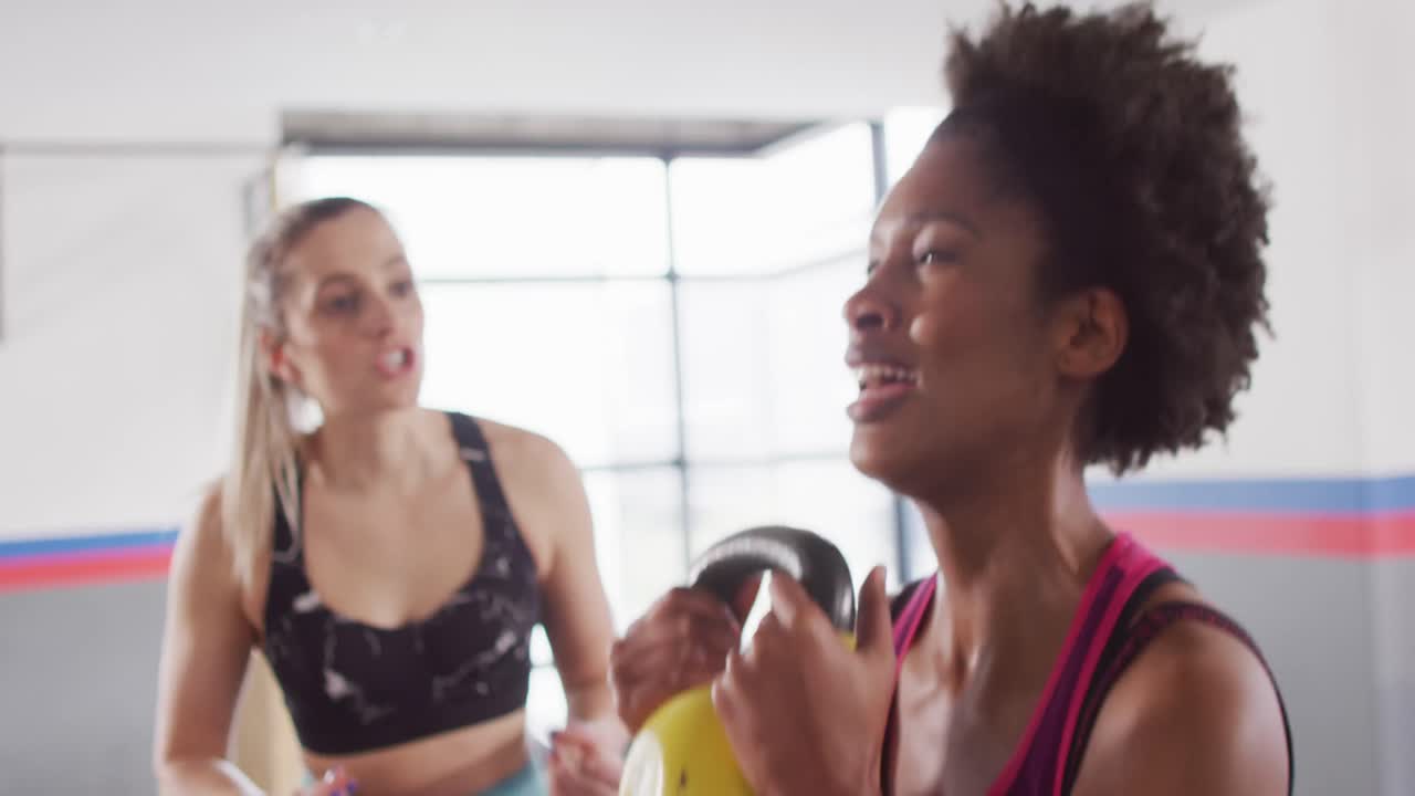 video de una entrenadora de fitness femenina diversa alentando a una mujer a hacer sentadillas con peso de kettlebell en el gimnasio