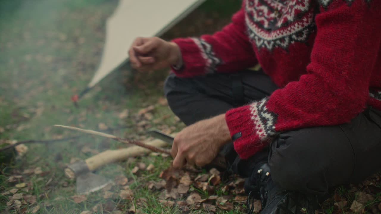 Close-up van man die met een mes een stok snijdt bij een kampvuur in een herfstbos