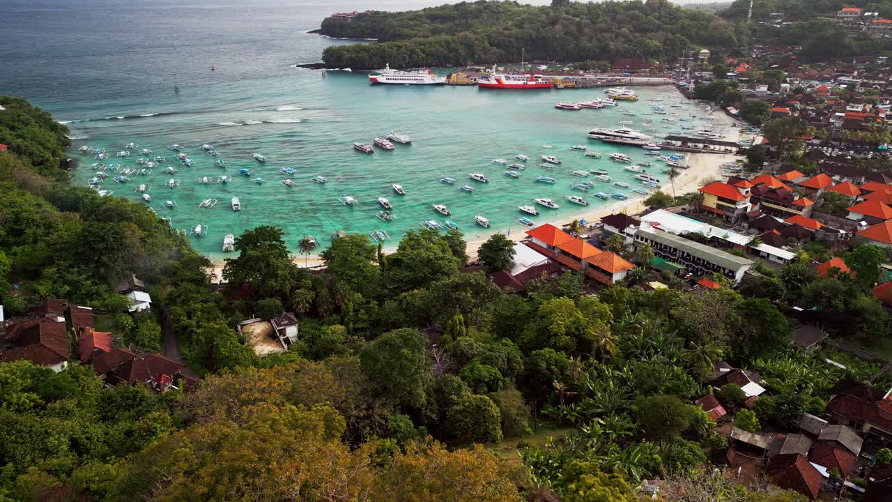 Aerial drone footage capturing Padang Bai Harbor in Karangasem, Bali. The turquoise water, fishing boats, and Blue Lagoon create a stunning coastal scene surrounded by tropical beauty