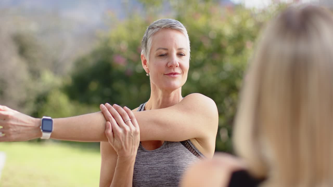 Instructora de fitness guiando a una amiga en un estiramiento de hombros en el patio, usando un reloj inteligente.
