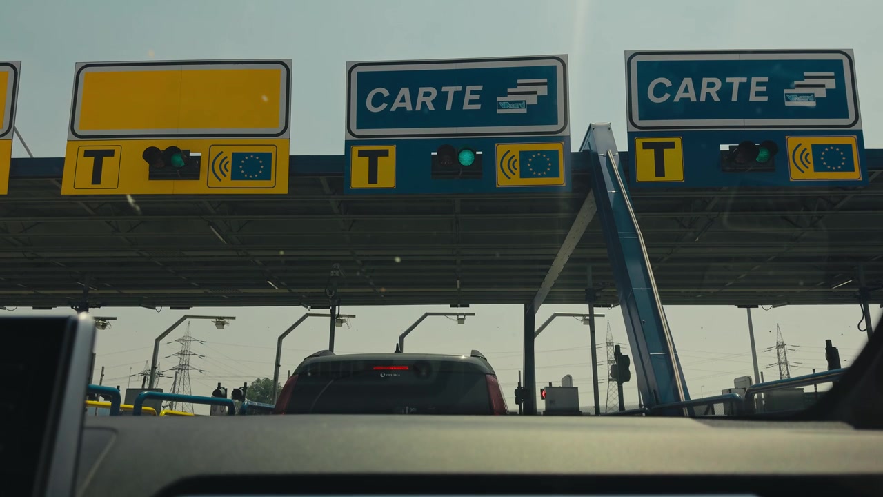Vista em primeira pessoa de um carro aproximando-se de uma praça de pedágio europeia com sinais de pagamento com cartão, semáforos e uma bandeira da UE, capturando a infraestrutura rodoviária moderna e as viagens