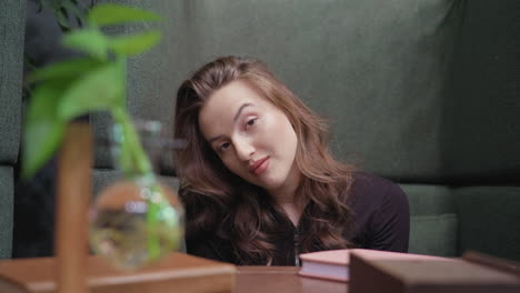 elegant woman with long wavy brown hair in black top slightly bends head and smiles at camera while seated beside wooden table with plant in glass vase, conveying calm relaxed expression