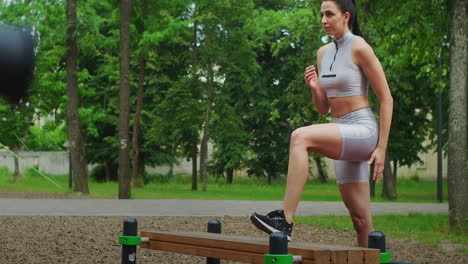 A-young-woman-in-a-Park-performs-striding-lunges-on-a-bench-in-sportswear-in-the-summer.-Athletics-Caucasian-woman-trains-in-the-Park.