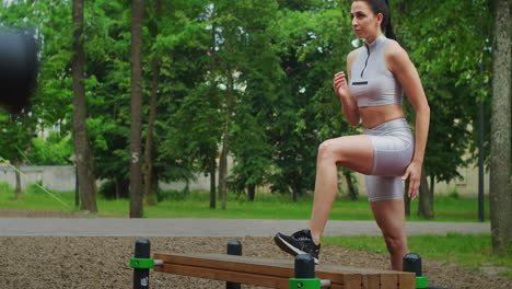 una mujer joven en un parque realiza estocadas en un banco en ropa deportiva en el verano. atletismo mujer caucásica entrena en el parque.