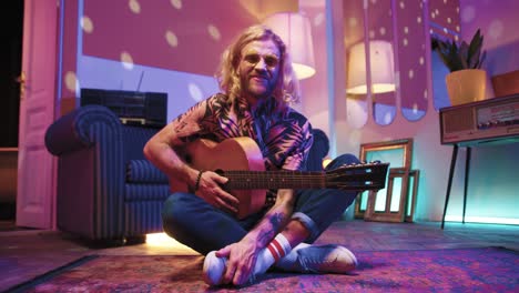 young stylish caucasian man with long hair sitting on the floor and playing the guitar in a retro party at home