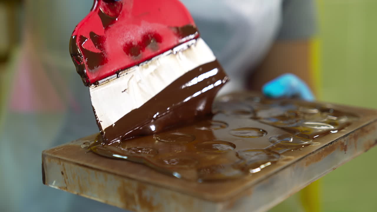 Chocolatier filling the molds with melted black chocolate. Candy maker uses spatula to spread chocolate over the molds.