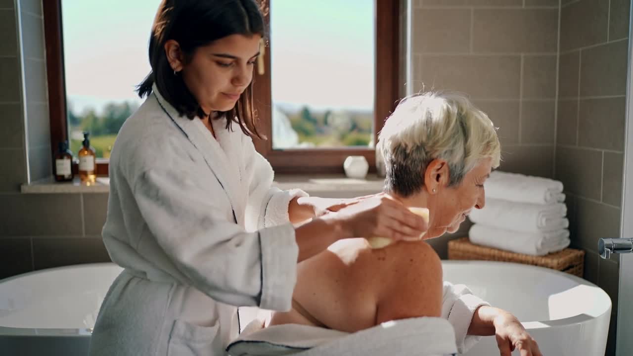 Ein Betreuer im Bademantel reinigt mit einem Schwamm den oberen Rücken eines Senioren im Badezimmer zum Baden mit Wanne.