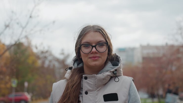 Close up of lady wearing glasses walking and smiling as wind blows hair across face with blurred background featuring passerby, moving cars, trees with autumn leaves, and buildings