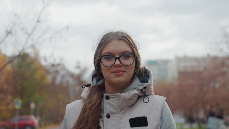 close up of lady wearing glasses walking and smiling as wind blows hair across face with blurred background featuring passerby, moving cars, trees with autumn leaves, and buildings
