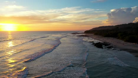 Vuelo-Aéreo-Desde-La-Playa-Hasta-El-Atardecer-Reflejándose-En-El-Agua-Con-Olas-Y-Surfistas-En-El-Océano-Pacífico-En-Tamarindo,-Costa-Rica