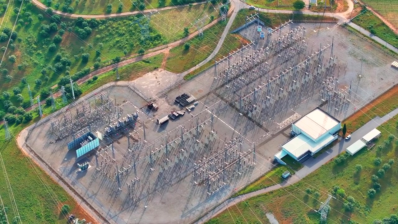 Aerial view of a high voltage power substation shows transmission lines and energy distribution equipment. The electrical grid is visible with nearby infrastructure and facilities.