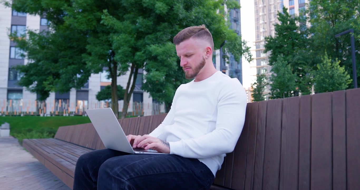 Young Man Working on Laptop on a Bench in an Urban Park