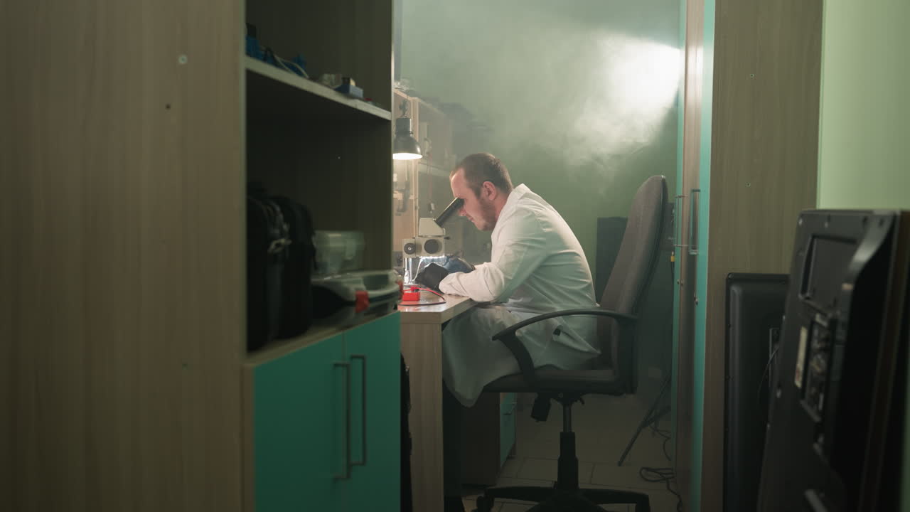 una vista de cerca de un técnico en una bata de laboratorio blanca trabajando diligentemente en componentes electrónicos bajo un microscopio con la luz encendida detrás de él y un estante junto a la entrada