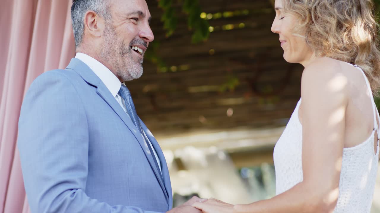 Coppia che si tiene per mano mentre la telecamera arretra sotto un pergolato a un matrimonio, uomo in abito e donna in vestito.