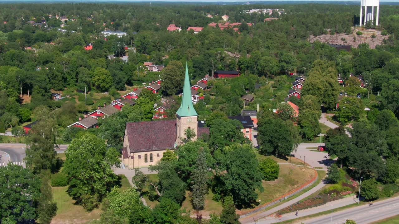 Splendida vista aerea di una chiesa tradizionale con guglia verde circondata da case svedesi rosse e una lussureggiante foresta a Gustavsberg